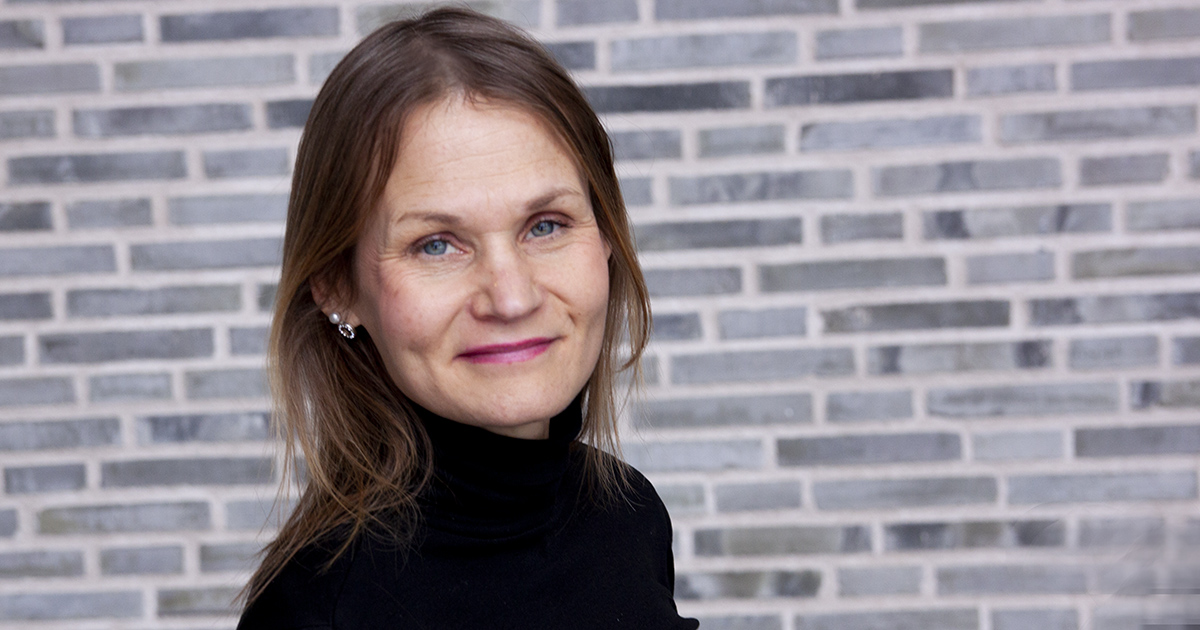 Kvinna med långt brunt hår i svart tröja ler mot kameran framför en tegelvägg.