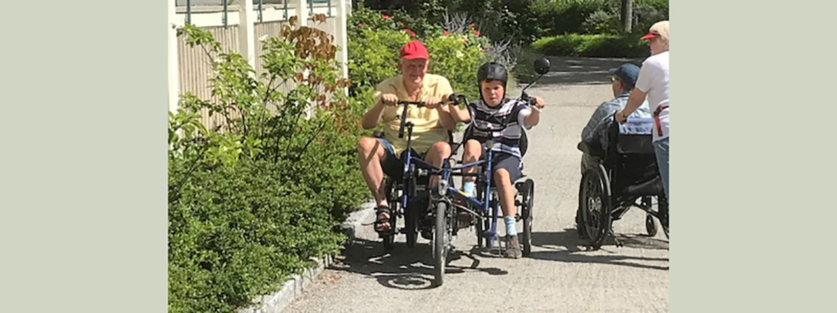 Två personer cyklar på tandemcykel, och två andra personer rullar fram i rullstol på en solig gångväg.