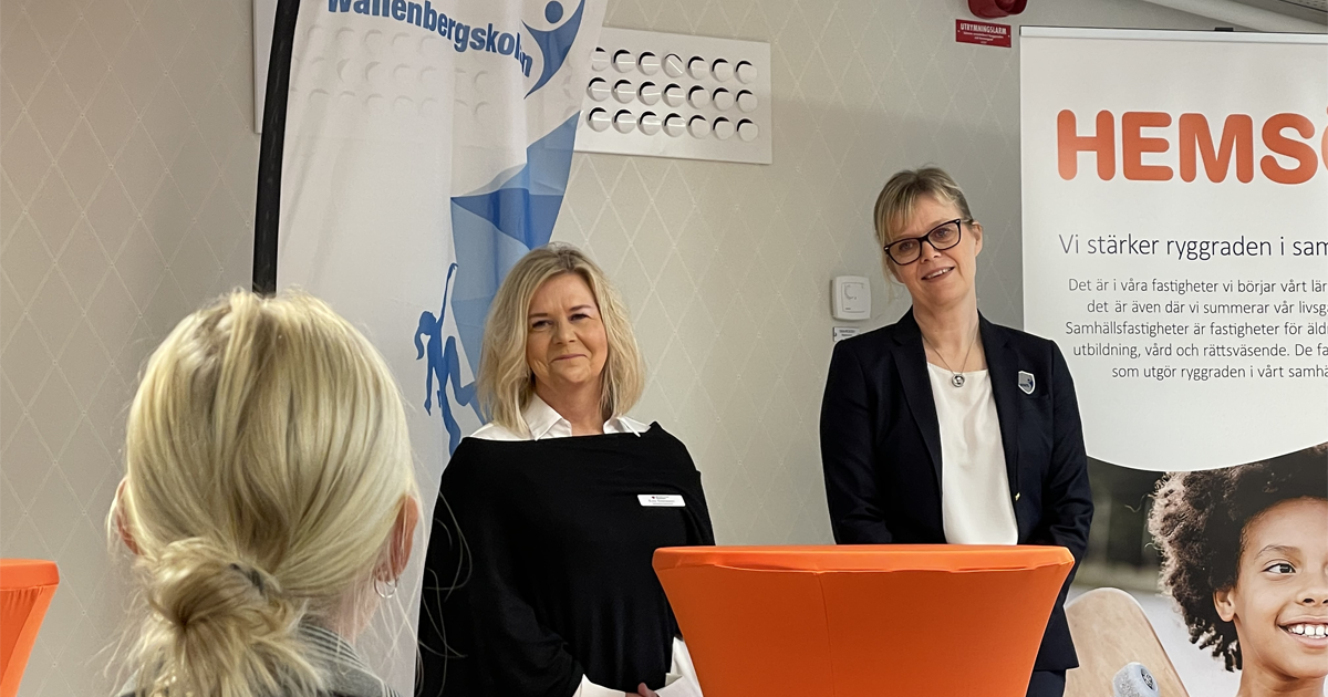 Två kvinnor står framför rollups, en åhörare syns bakifrån. Orange bord i förgrunden, företagsloggor i bakgrunden.