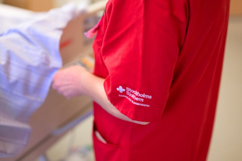Person i röd vårduniform med texten "Stockholms Sjukhem" bredvid en patientsäng.