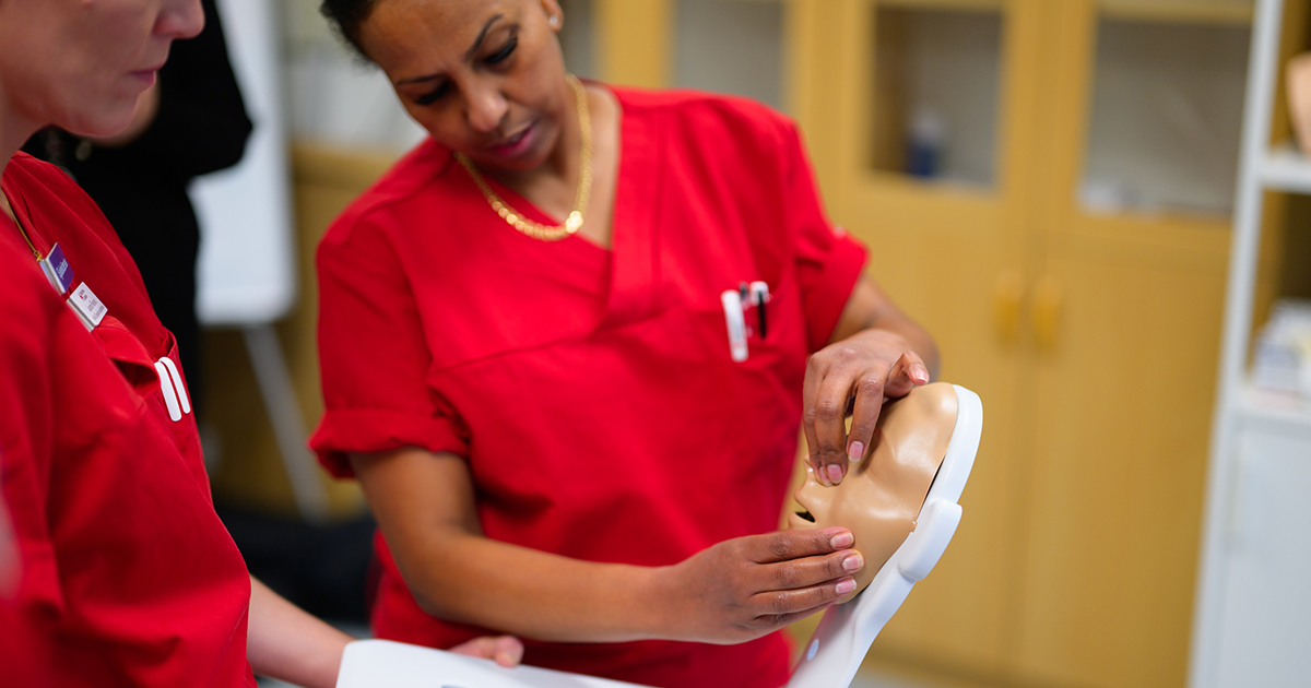 Två personer i röda sjukvårdskläder övar på en medicinsk modell av ett ansikte i ett klassrum.