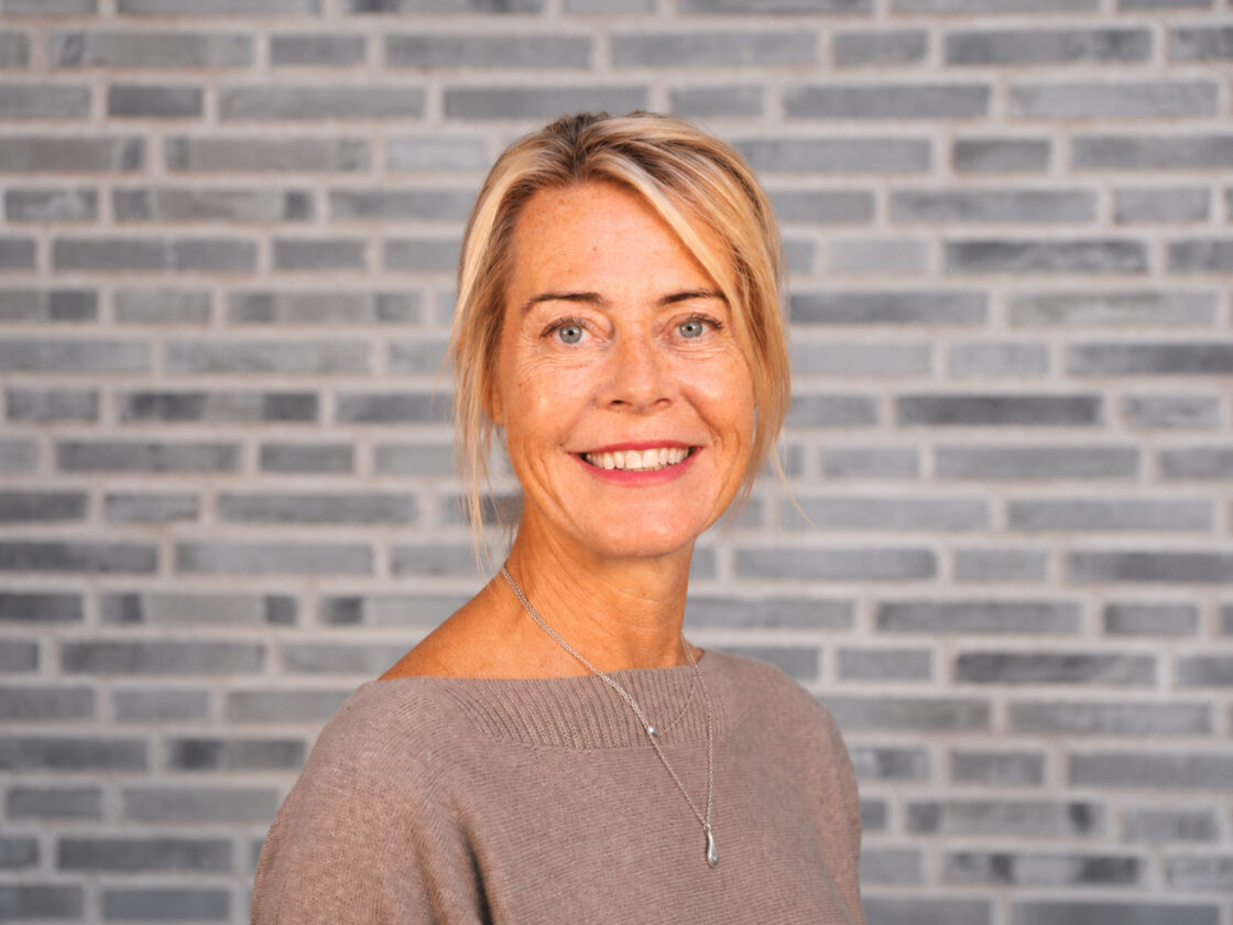 Leende kvinna med blont hår och beige tröja framför en vägg av grå tegelstenar.