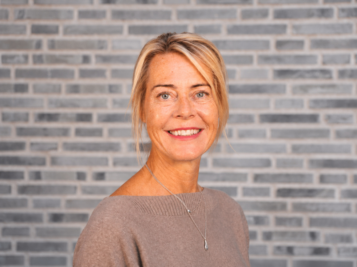 Leende kvinna med blont hår och beige tröja framför en vägg av grå tegelstenar.