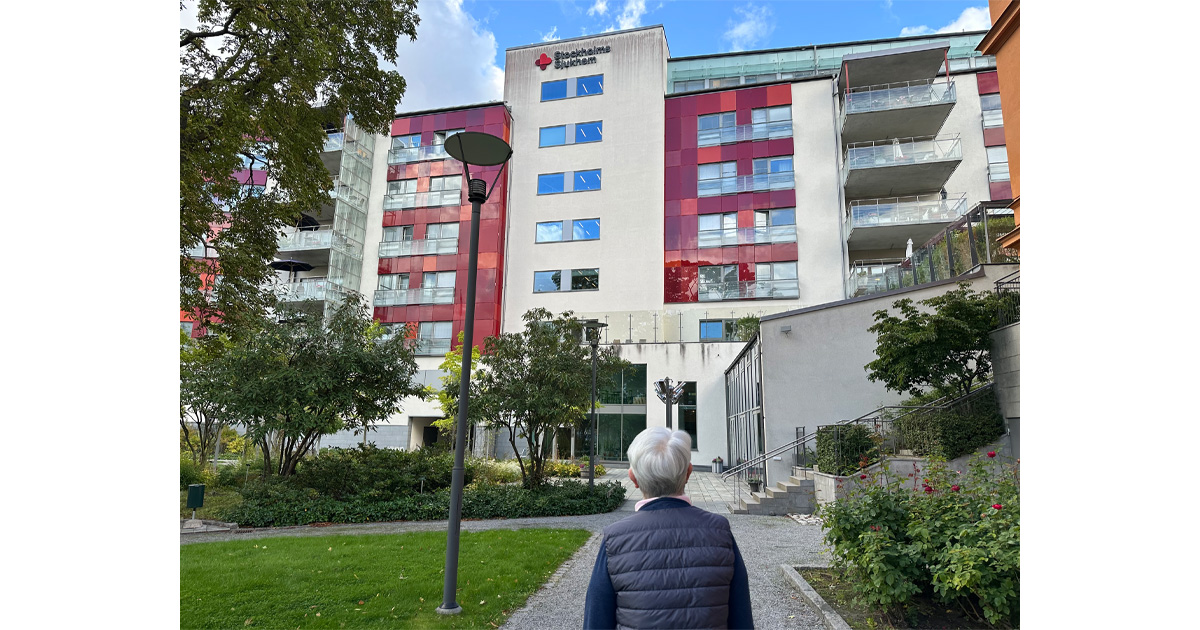 En äldre person står framför ett modernt sjukhus med rödvita fasader och grönskande trädgård.